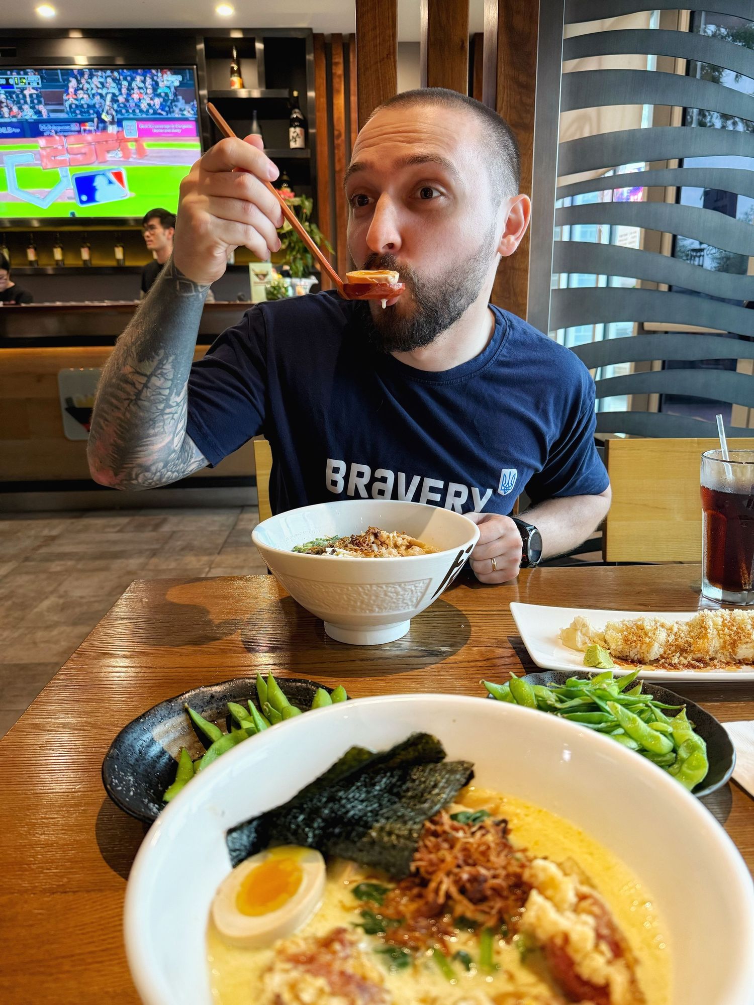 Diner enjoying ramen noodle soup with egg and nori while watching sports at a restaurant with a green salad on the side.