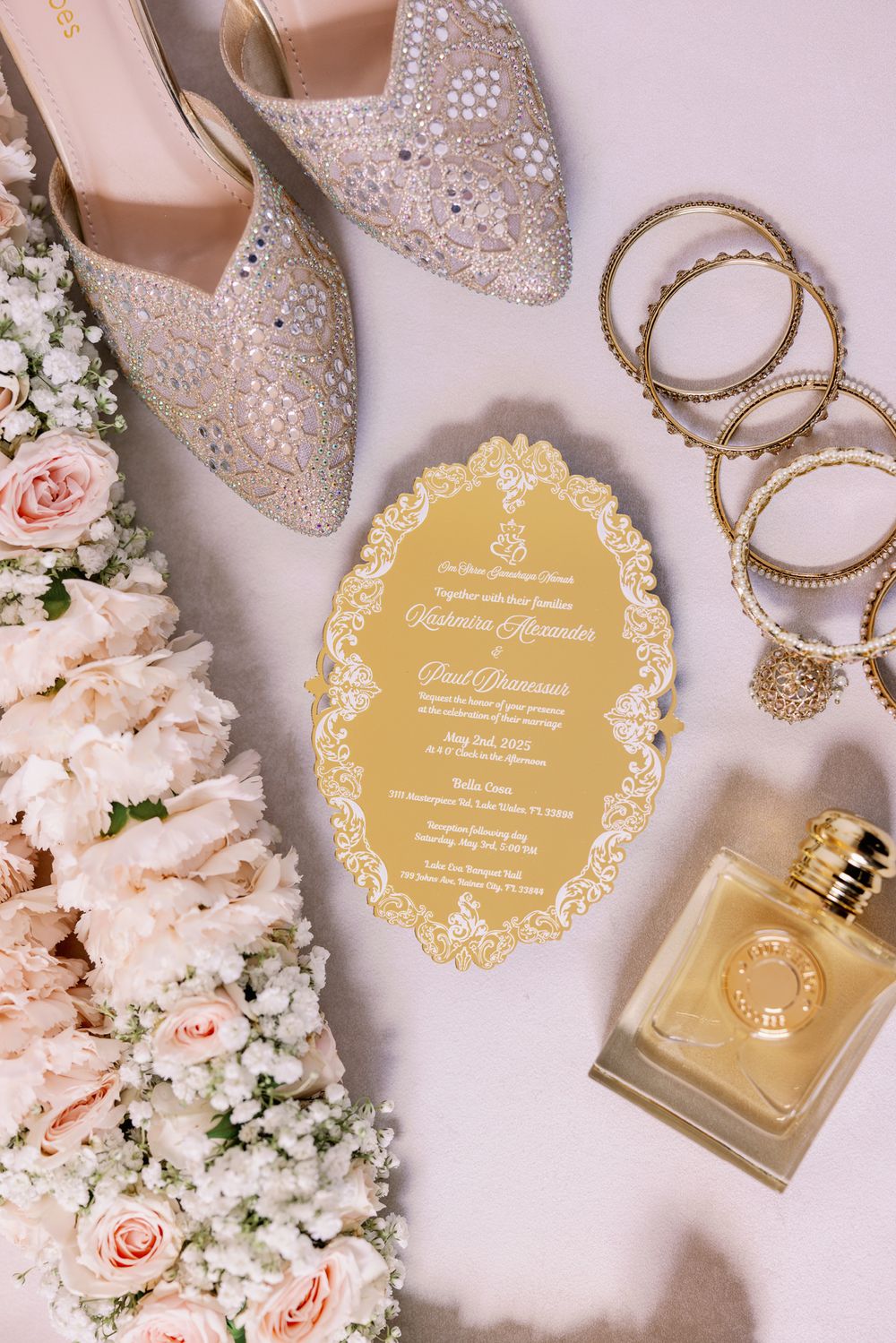 Elegant wedding invitation with sparkly shoes, perfume, gold bangles and blush roses arranged on light background.