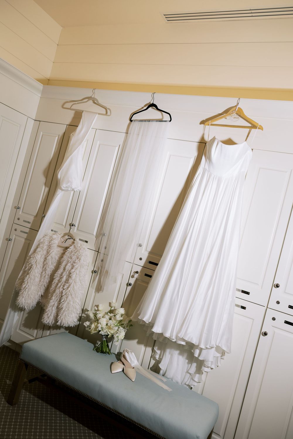 White wedding dresses and fur shawl hanging on hangers in a dressing room with built-in closets. NC WEDDING PHOTOGRAPHER, LUXURY WEDDING