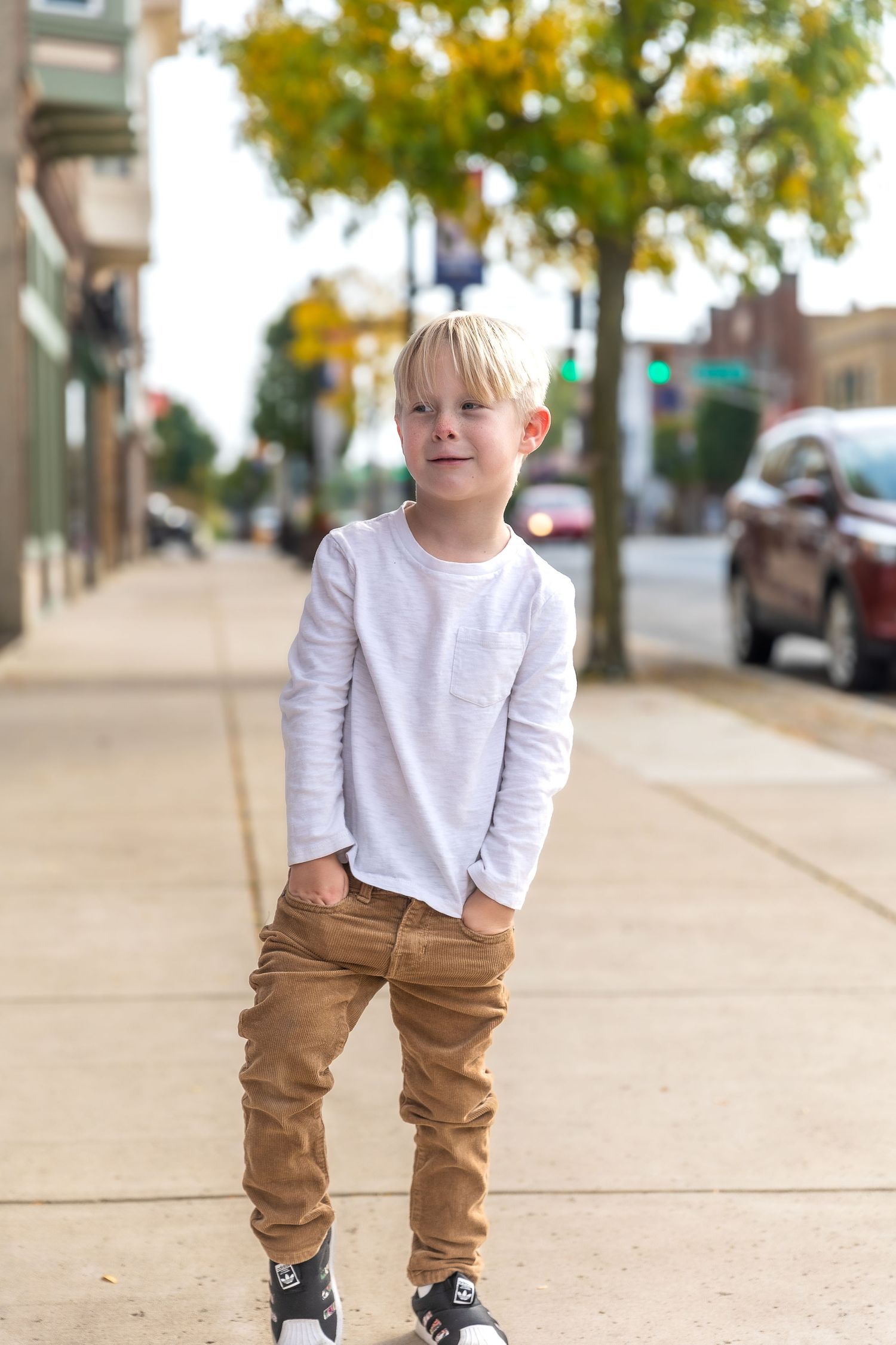 Young child in white long sleeve shirt and brown pants standing on city sidewalk with autumn trees in background.