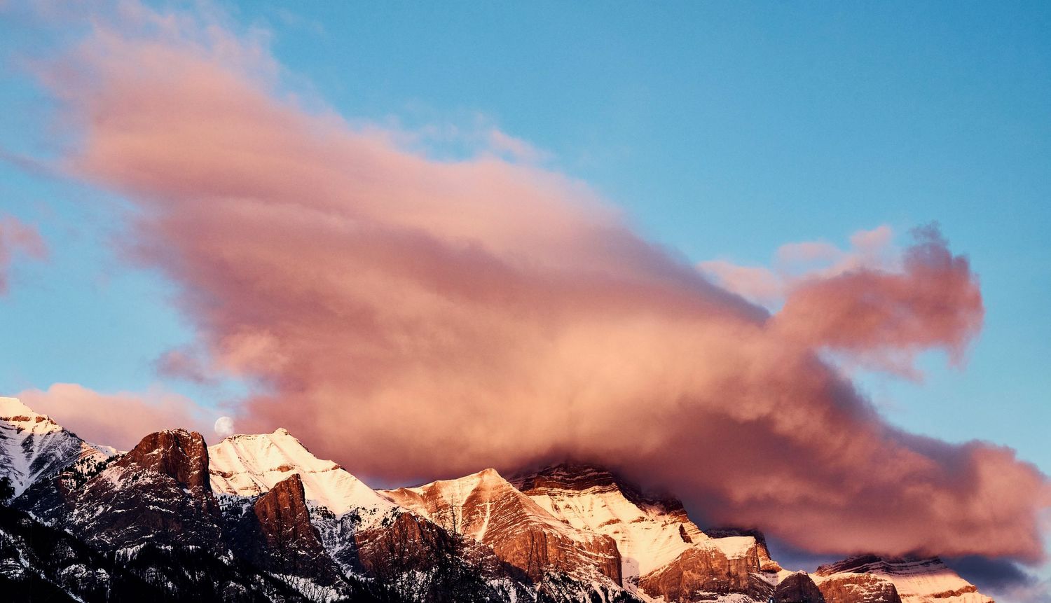 Pink sunset clouds drift over snow-capped Mount Rundle peaks against a vibrant blue sky in Canmore looking towards Banff