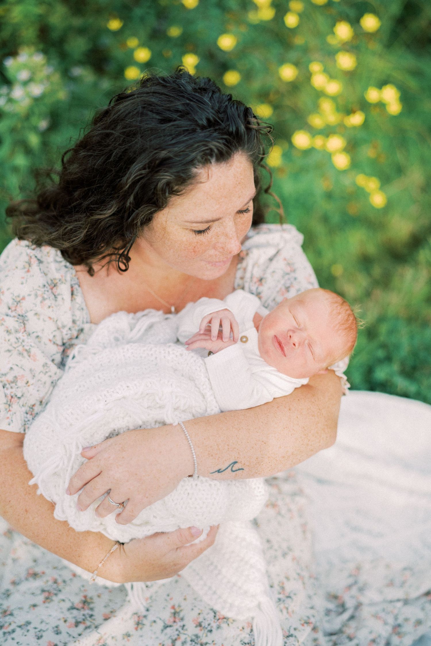 A tender outdoor portrait shows a mother cradling her newborn baby wrapped in white lace against a sunlit garden backdrop.