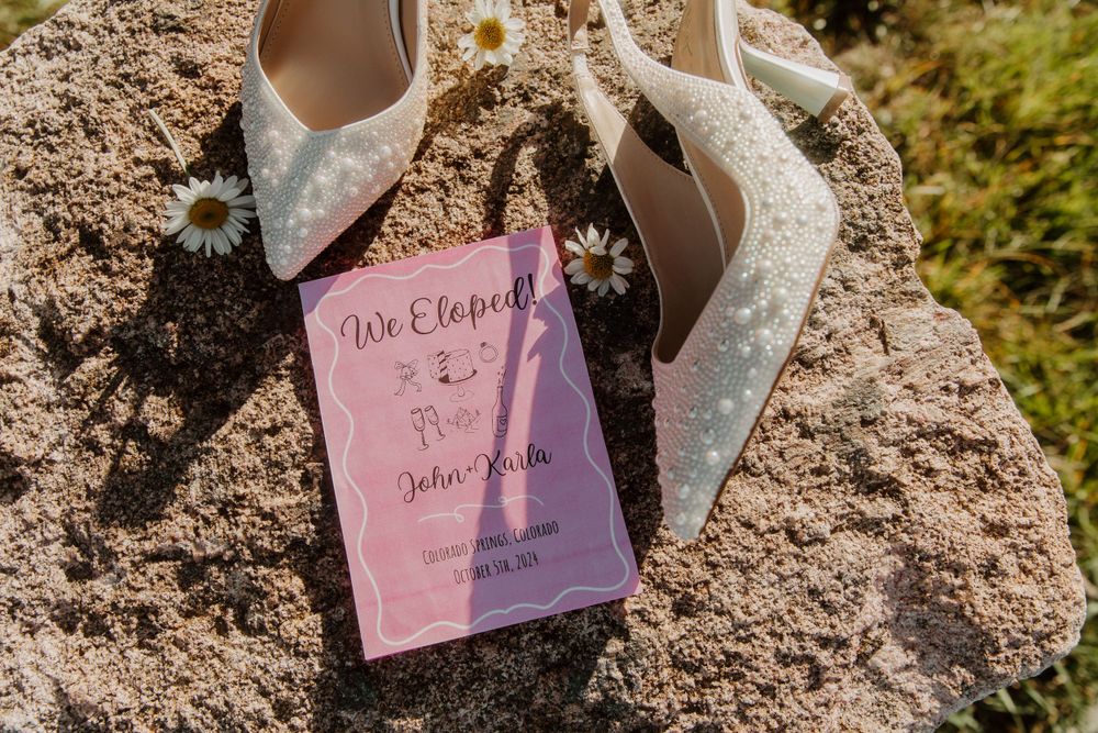 Wedding shoes and a pink invitation card displayed on a rocky surface in natural outdoor lighting in Colorado Springs, Colorado.