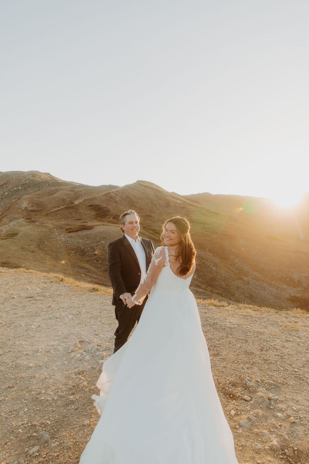 Couple in wedding attire stands on mountaintop during golden sunset in mountain landscape in Denver, Colorado.