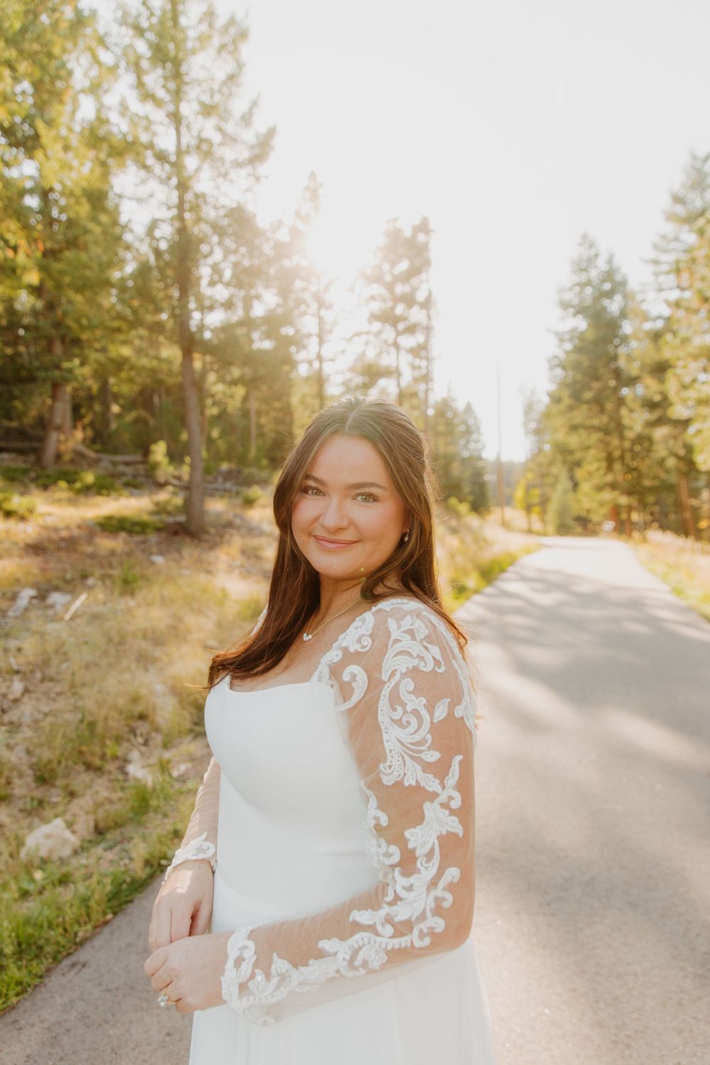 A bride in a white lace-sleeve wedding dress stands on a sunny forest road surrounded by tall pine trees in Denver, Colorado.