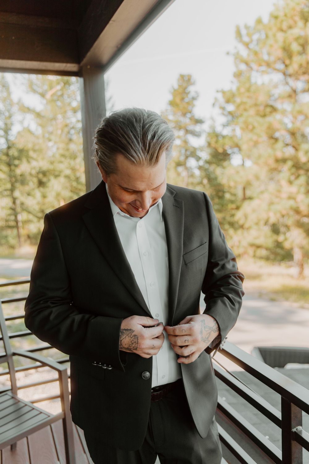 Well-dressed groom in dark suit buttons up jacket while standing on outdoor porch with pine trees in background in Denver, Colorado.