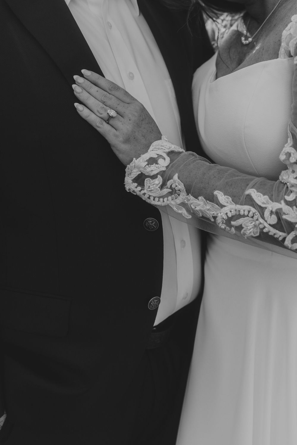 Close-up black and white shot of lace sleeve detail on wedding dress with engagement ring visible on hand.