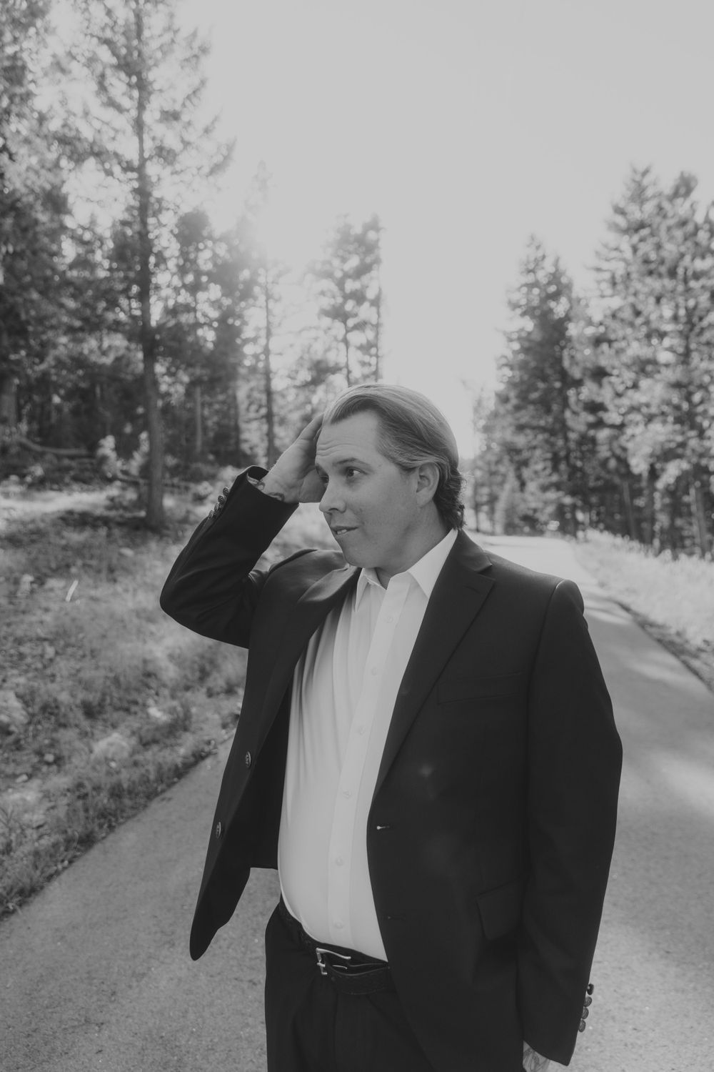 Black and white photo of person in formal attire standing on a forest road with tall pine trees in the background in Denver, Colorado.