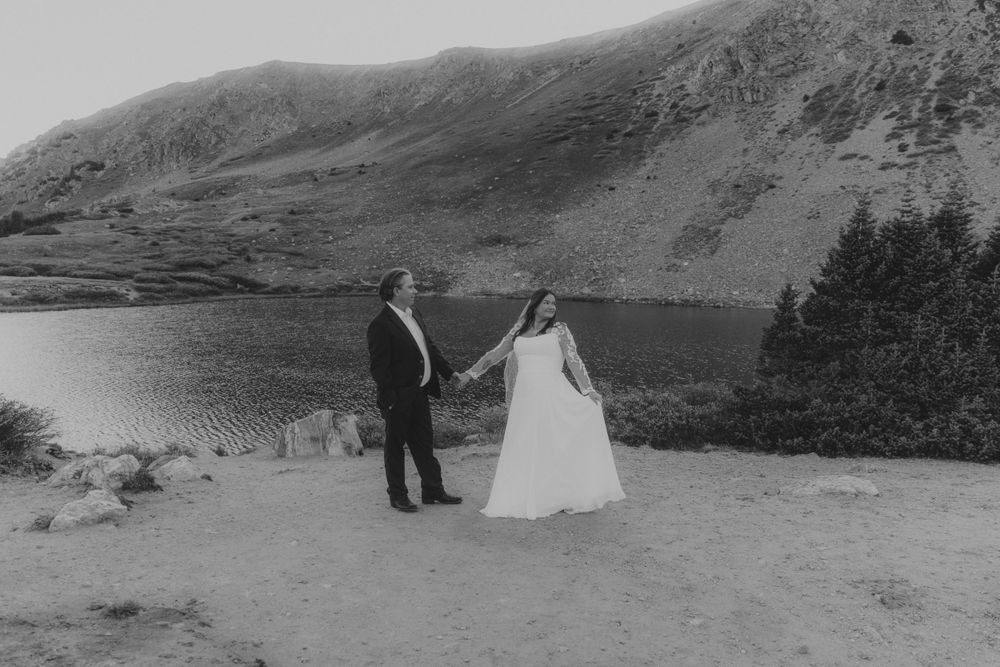A couple holds hands while standing by a serene mountainside lake in black and white photography in Denver, Colorado.
