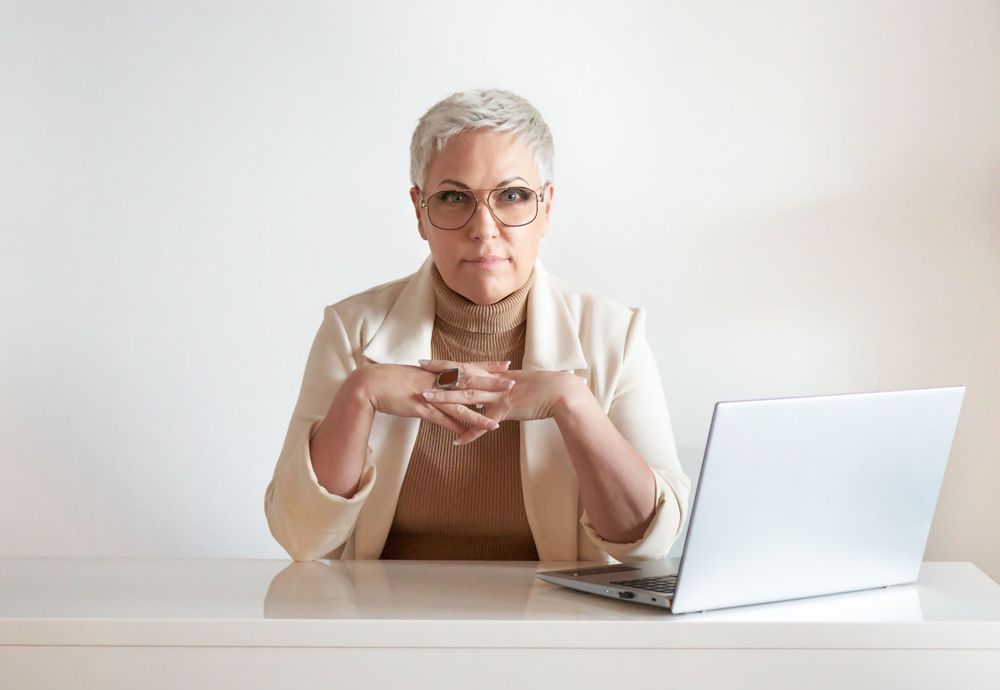 Denise Frenette in a white blazer and glasses sits at desk with laptop while gesturing during conversation photographed by Evan Will