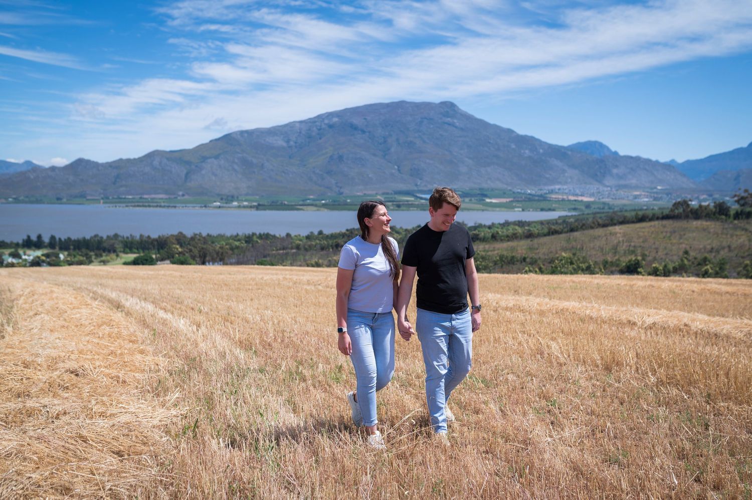 Two people walk hand in hand through a golden wheat field with mountains and blue skies in the background.