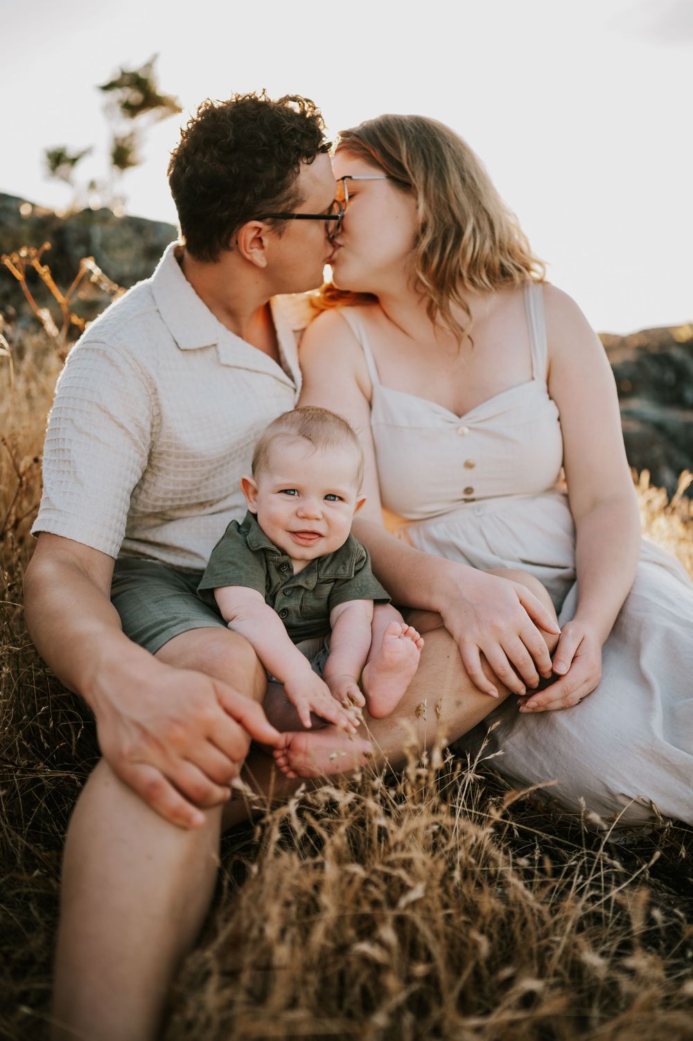 Photo of family sitting on rocky bluff grass kissing Neck Point Park Nanaimo sunset family photography by Vancouver Island photographer