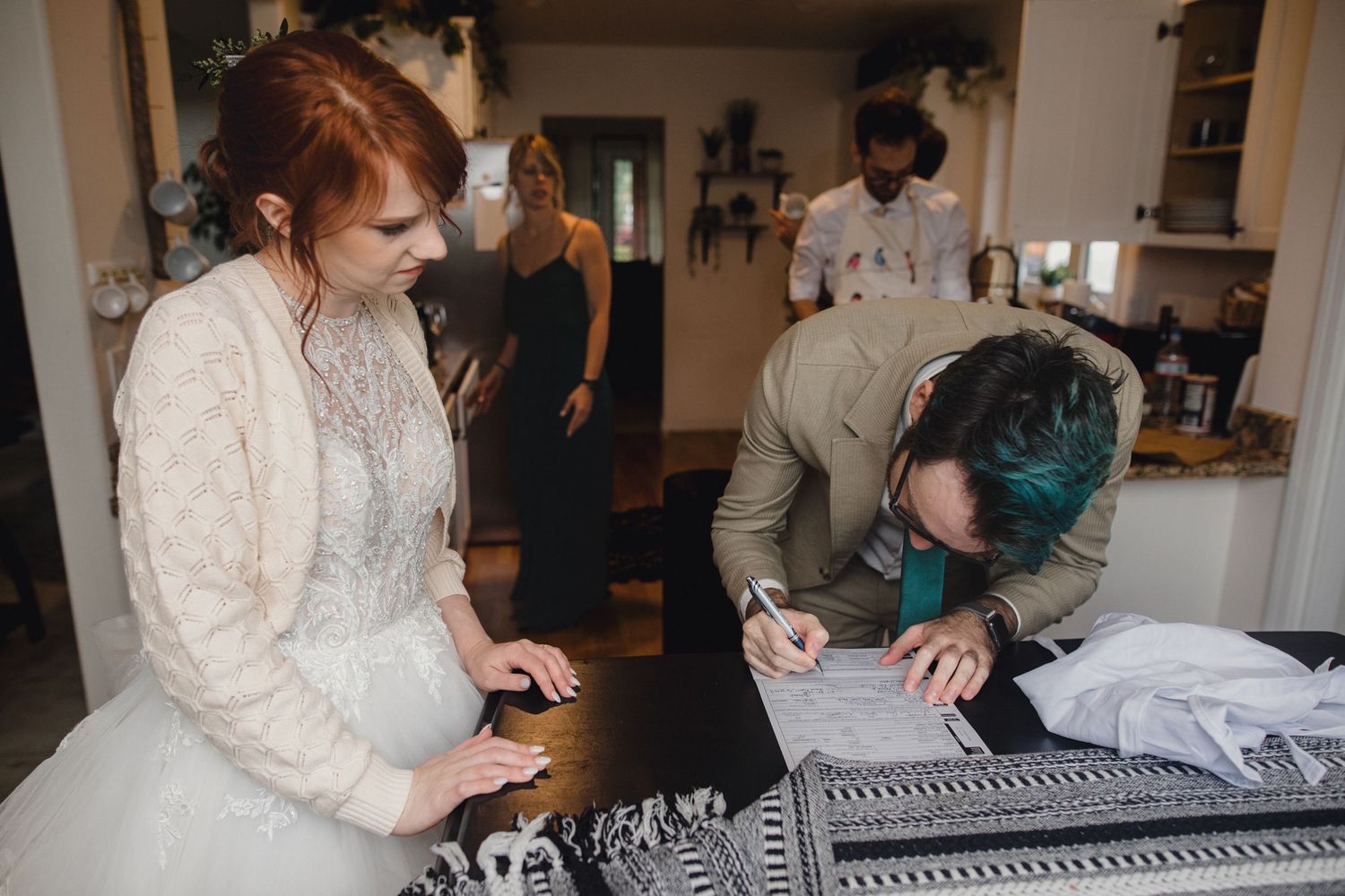 A person in a white wedding dress examines clothing details while another person looks down at a table in a kitchen setting.