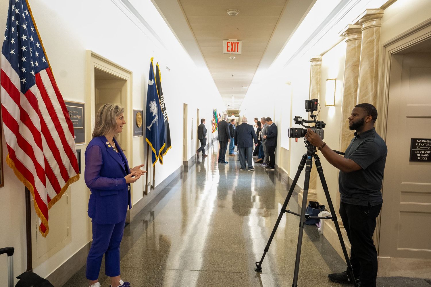 People stand in a government building hallway with US flags visible along the walls as cameras film.