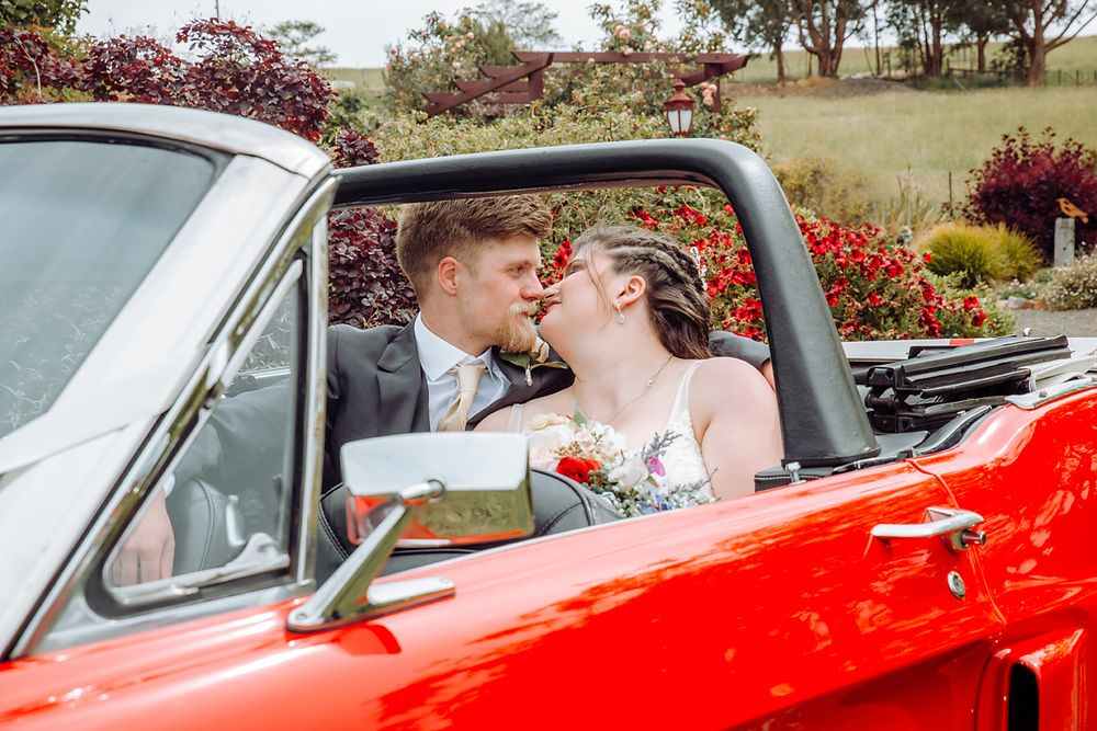 A romantic moment captured in a bright red vintage car surrounded by vibrant blooming flowers.