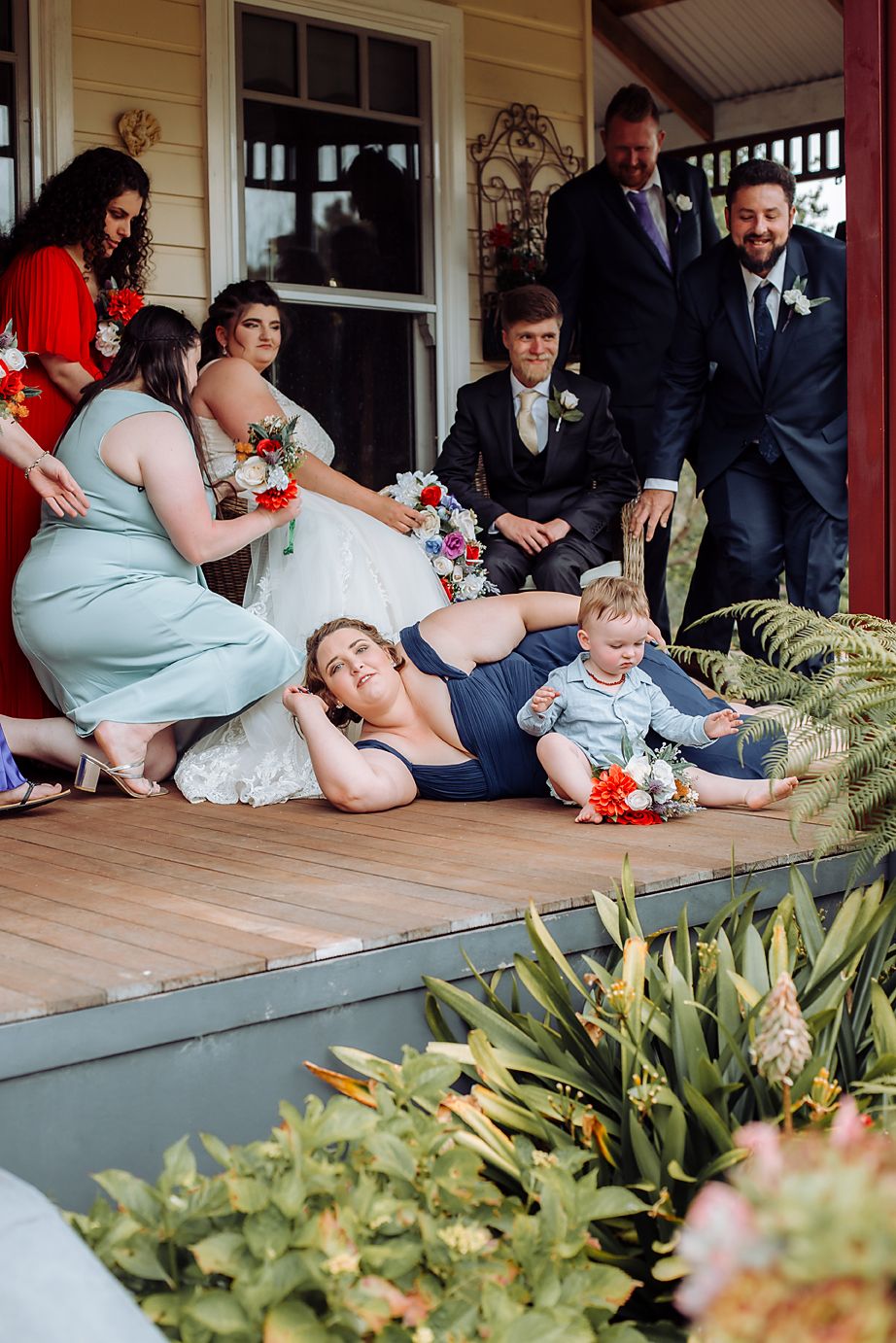 Several wedding party members sit together on wooden porch steps during outdoor photo session on sunny day.