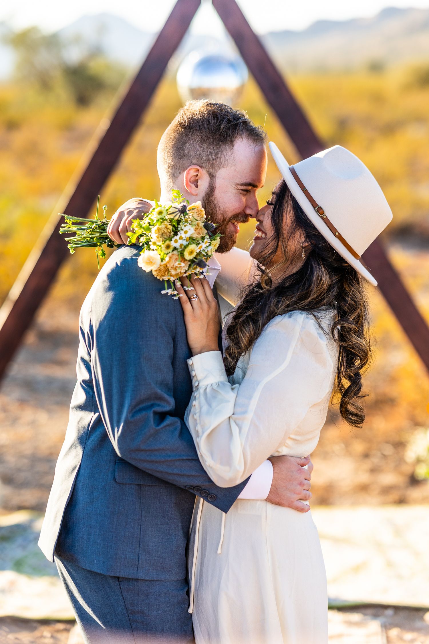 A romantic couple in wedding attire share an intimate moment at sunset with a wooden triangle arch and yellow flowers.