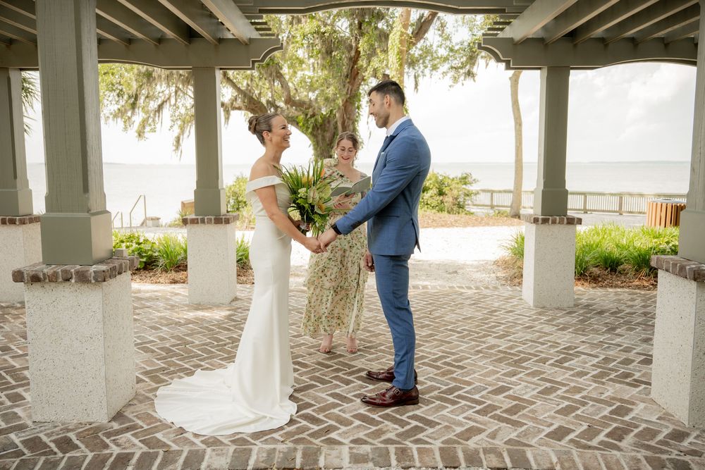 A wedding ceremony takes place under a covered brick patio with white columns overlooking palm trees and water.