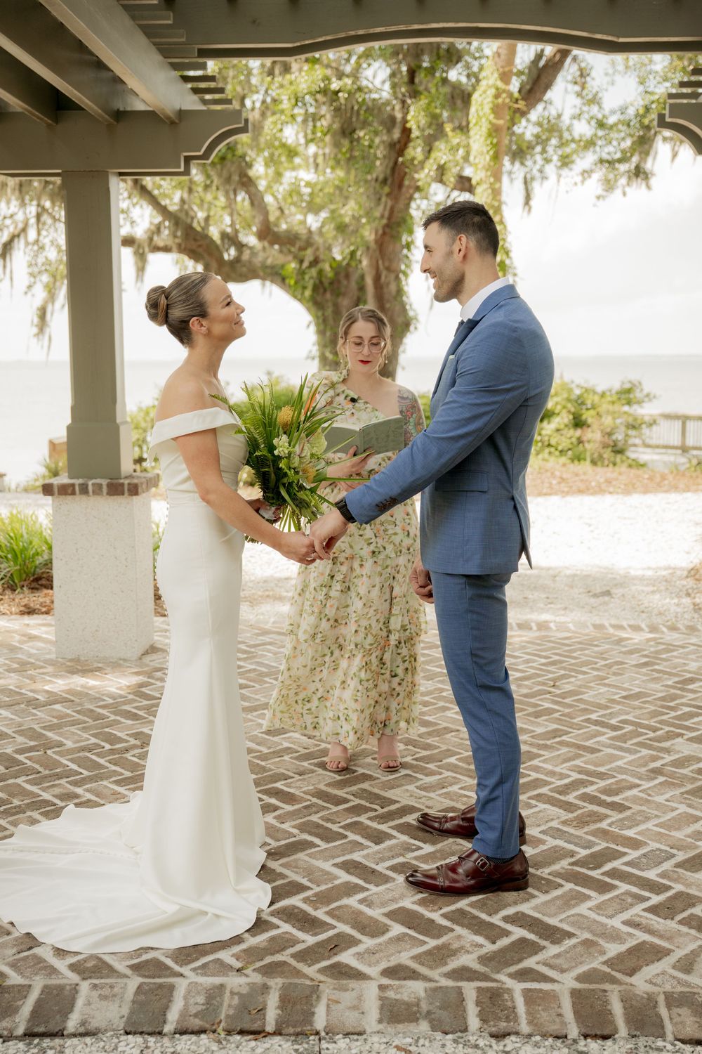 A couple exchanges wedding vows on a brick patio beneath a covered pavilion while an officiant stands between them.