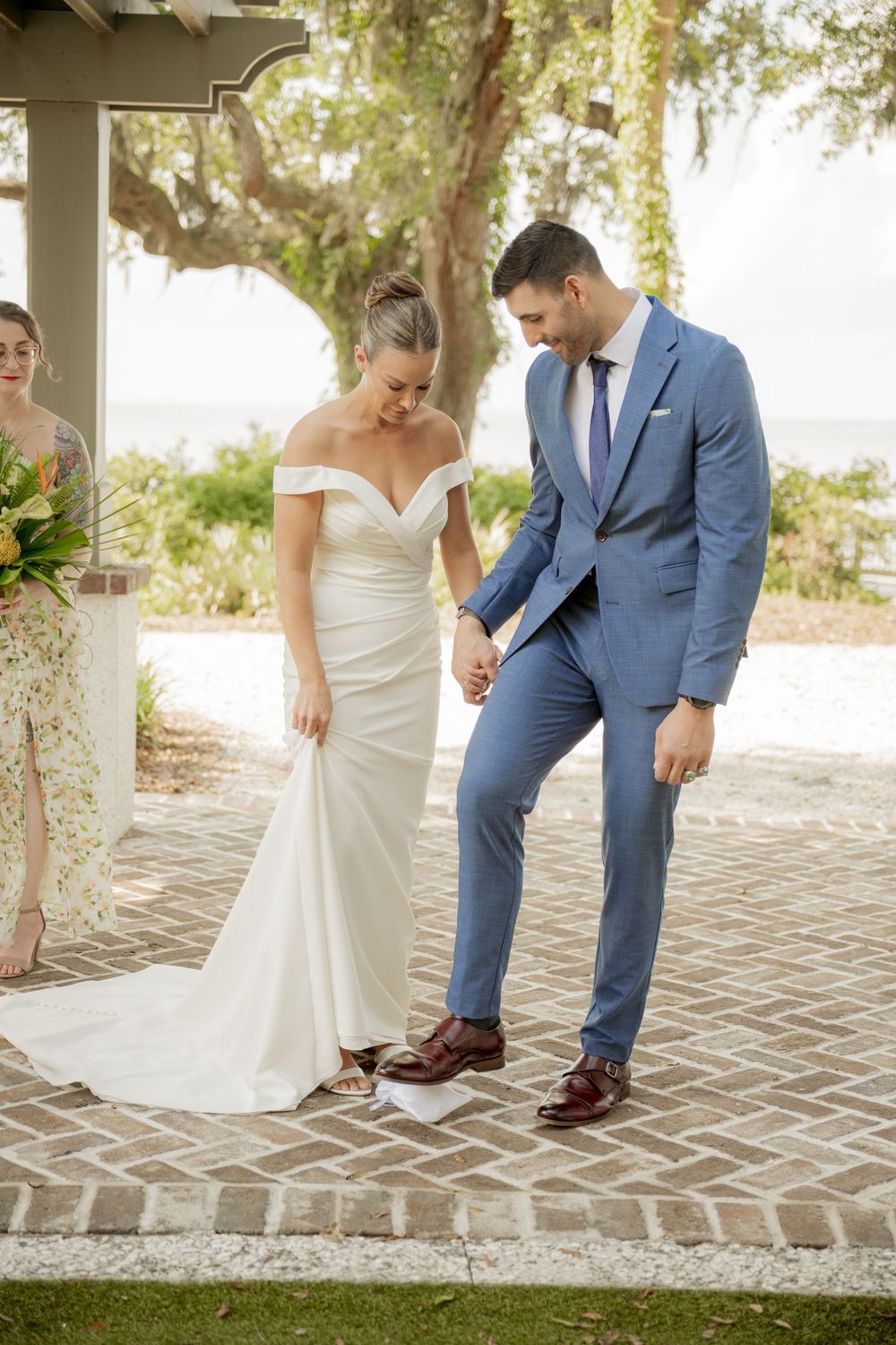 A couple in wedding attire admiring their footwear on a brick patio with lush trees in the background.