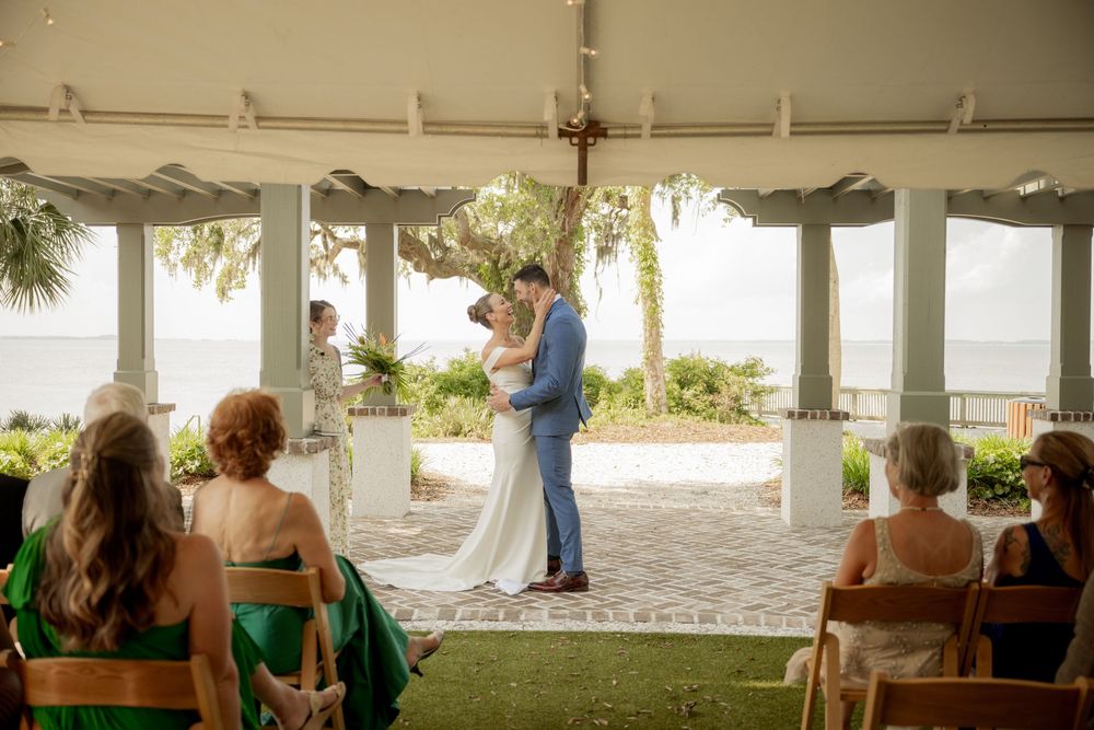 A wedding ceremony takes place under an open-air pavilion overlooking the water with guests seated on wooden chairs.