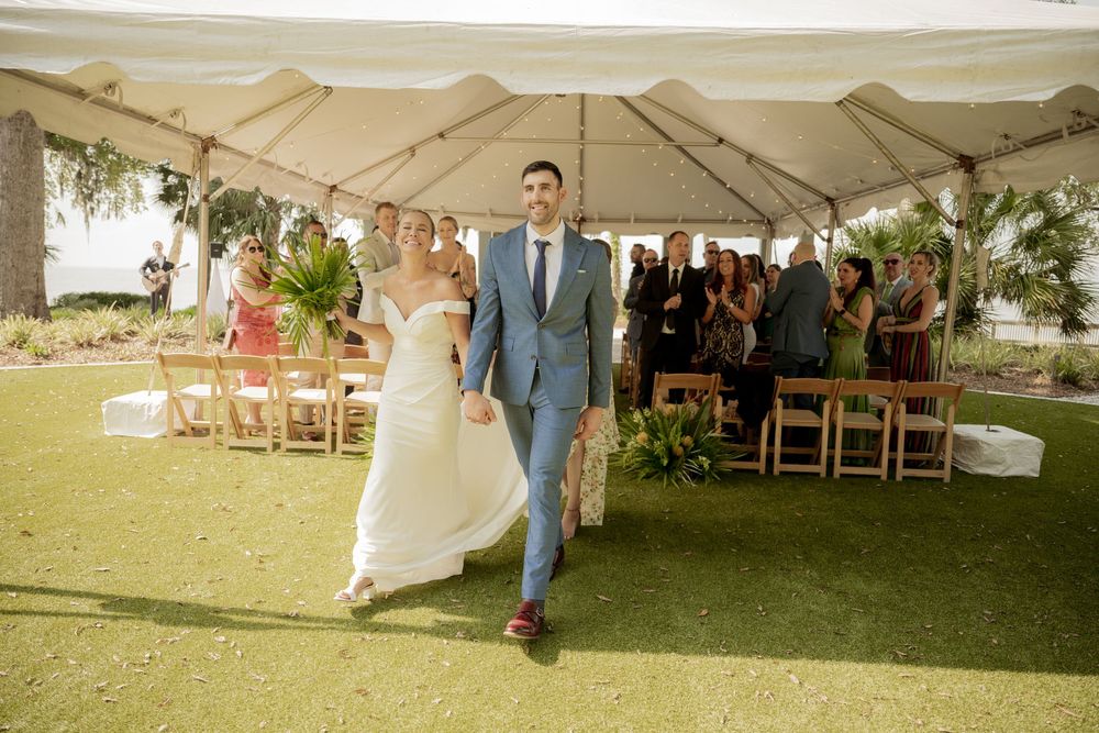 A newlywed couple joyfully exits their outdoor wedding ceremony under a white tent while guests look on.
