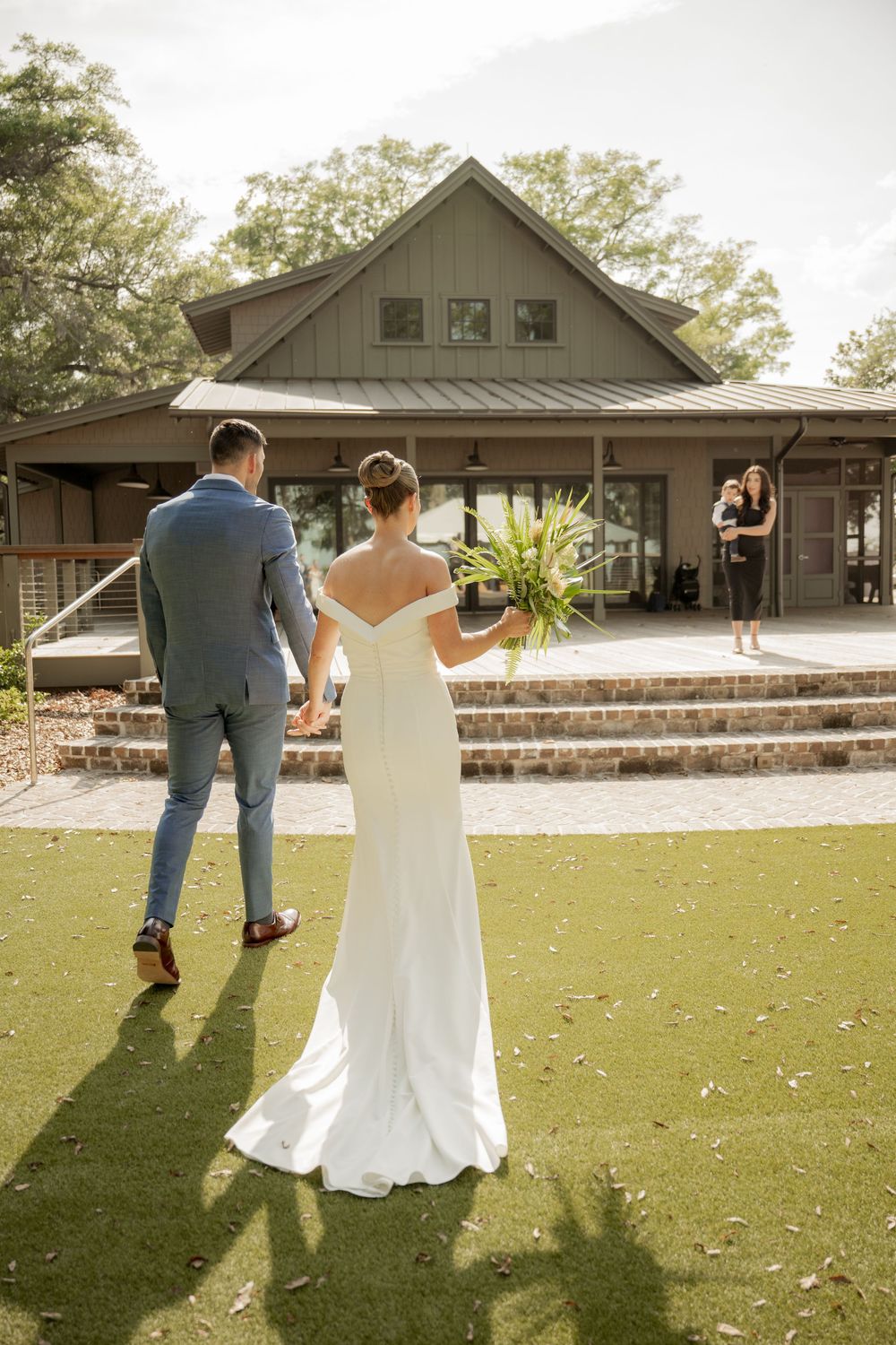 A couple in wedding attire walk towards a rustic lodge venue with wooden architecture and brick steps on a green lawn.