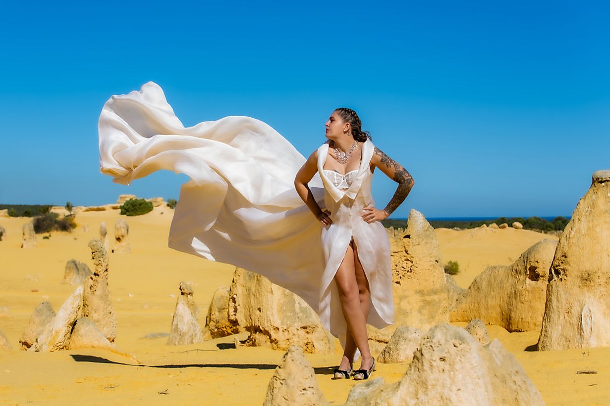 A flowing white dress billows dramatically against a bright blue sky at the Pinnacles Desert limestone formations in Australia.
