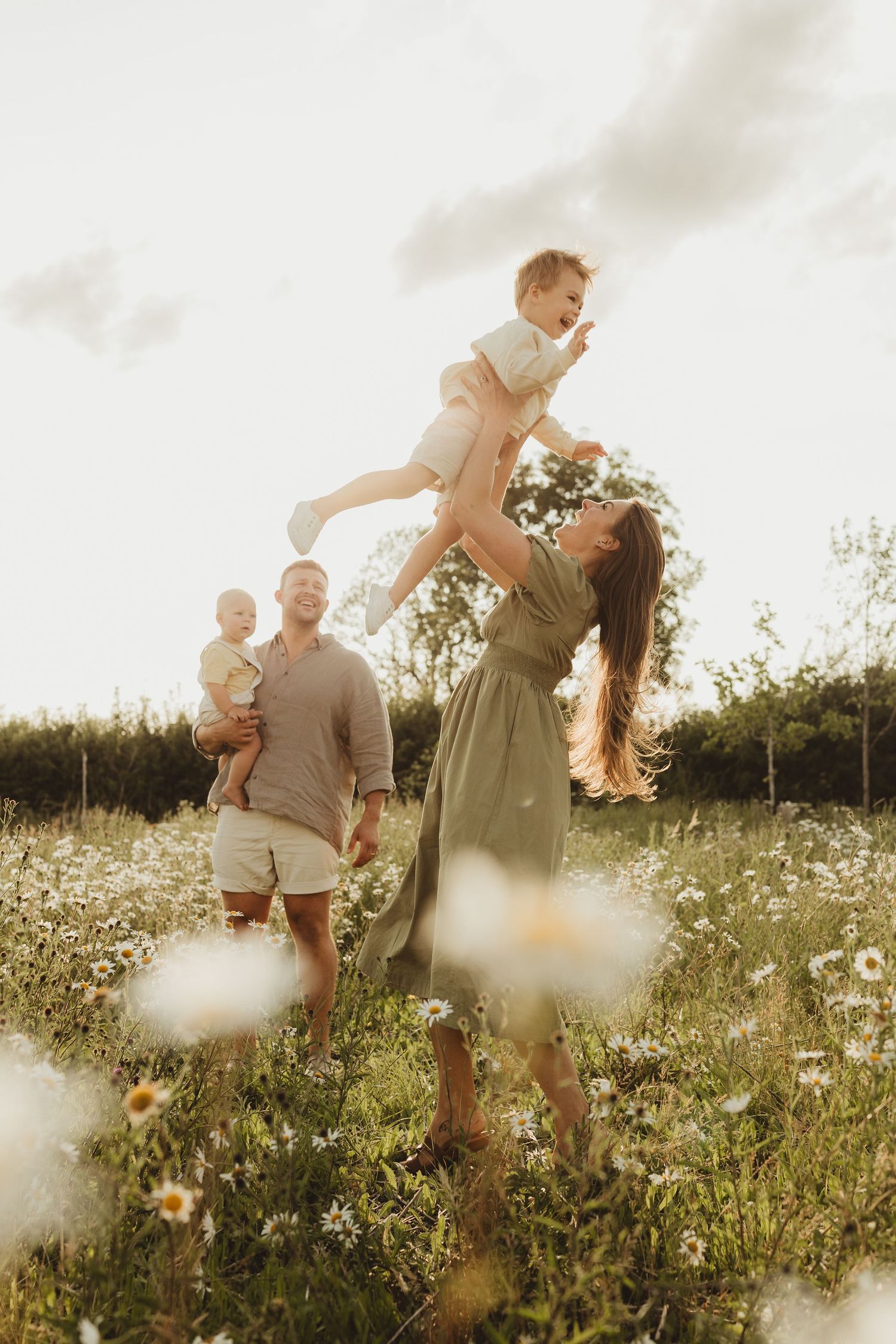 Family playing together in a sunlit wildflower meadow during golden hour while wearing coordinated neutral outfits.