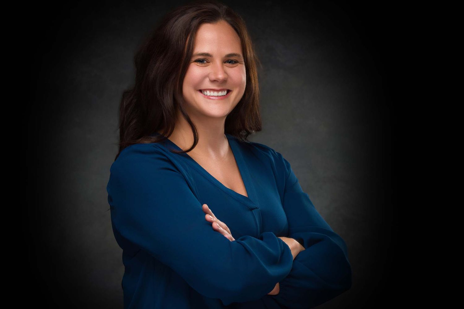 Professional headshot portrait of someone in a dark blue top against a black background with confident crossed arms pose.