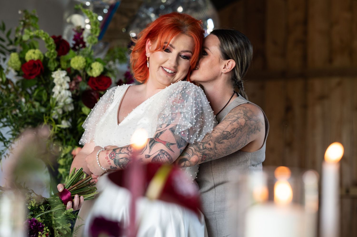 Two brides hug, they are framed by foliage and candles