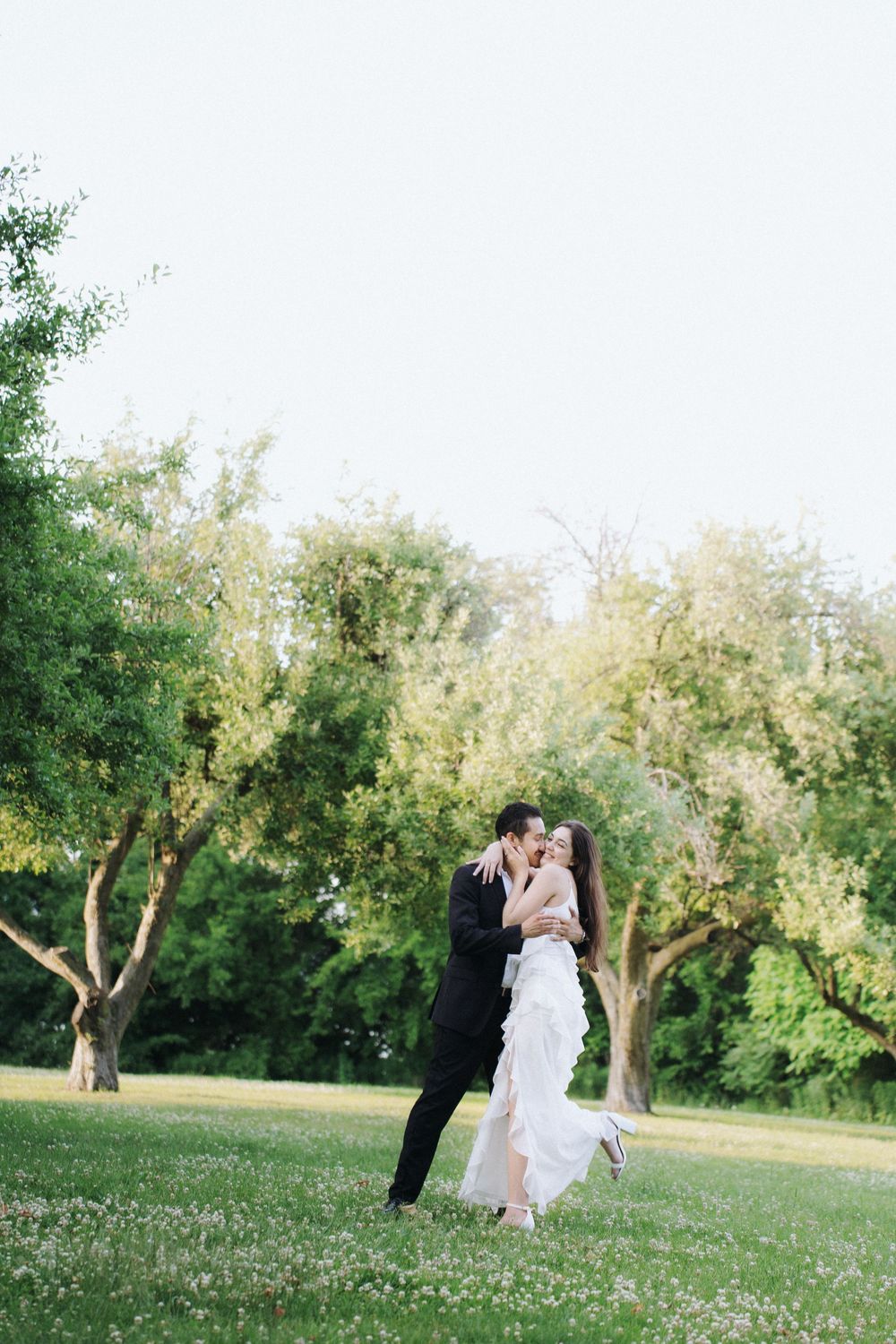 A couple in wedding attire embrace in a lush green garden setting with sunlit trees in the background.