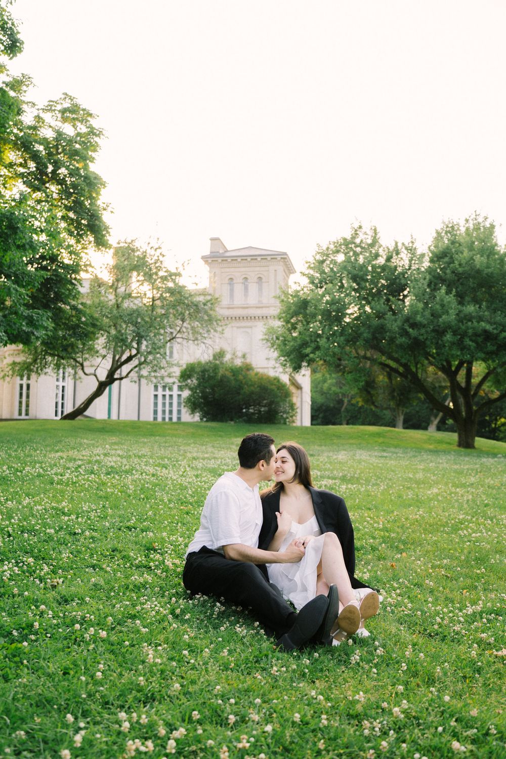 A couple enjoys a romantic moment on a grassy lawn in front of a grand historic building with white architecture.