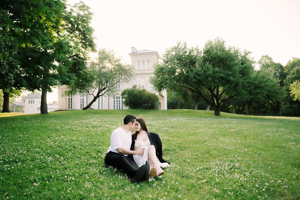A couple sits together on a lush green lawn surrounded by trees with a building visible in the background.