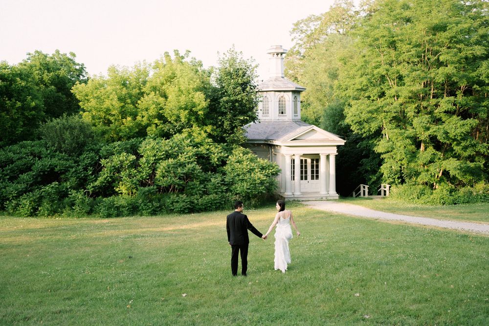Two people walking on a green lawn towards a white gazebo structure surrounded by lush trees on a sunny day.