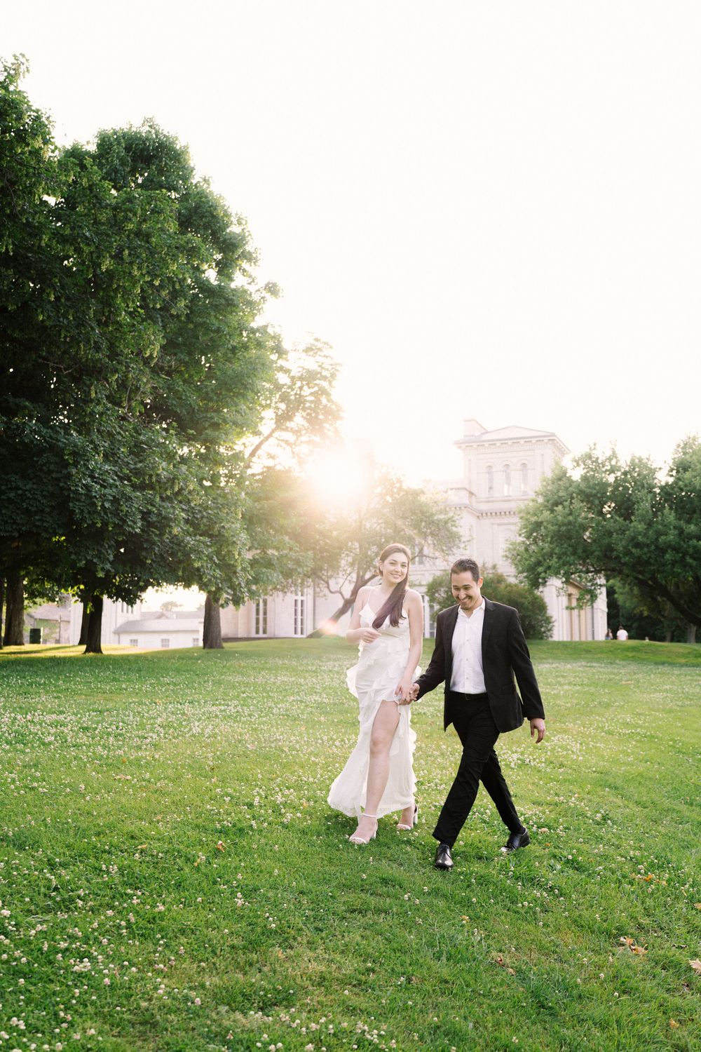 Two people walking together on a grassy lawn at sunset with a building visible in the background through the trees.