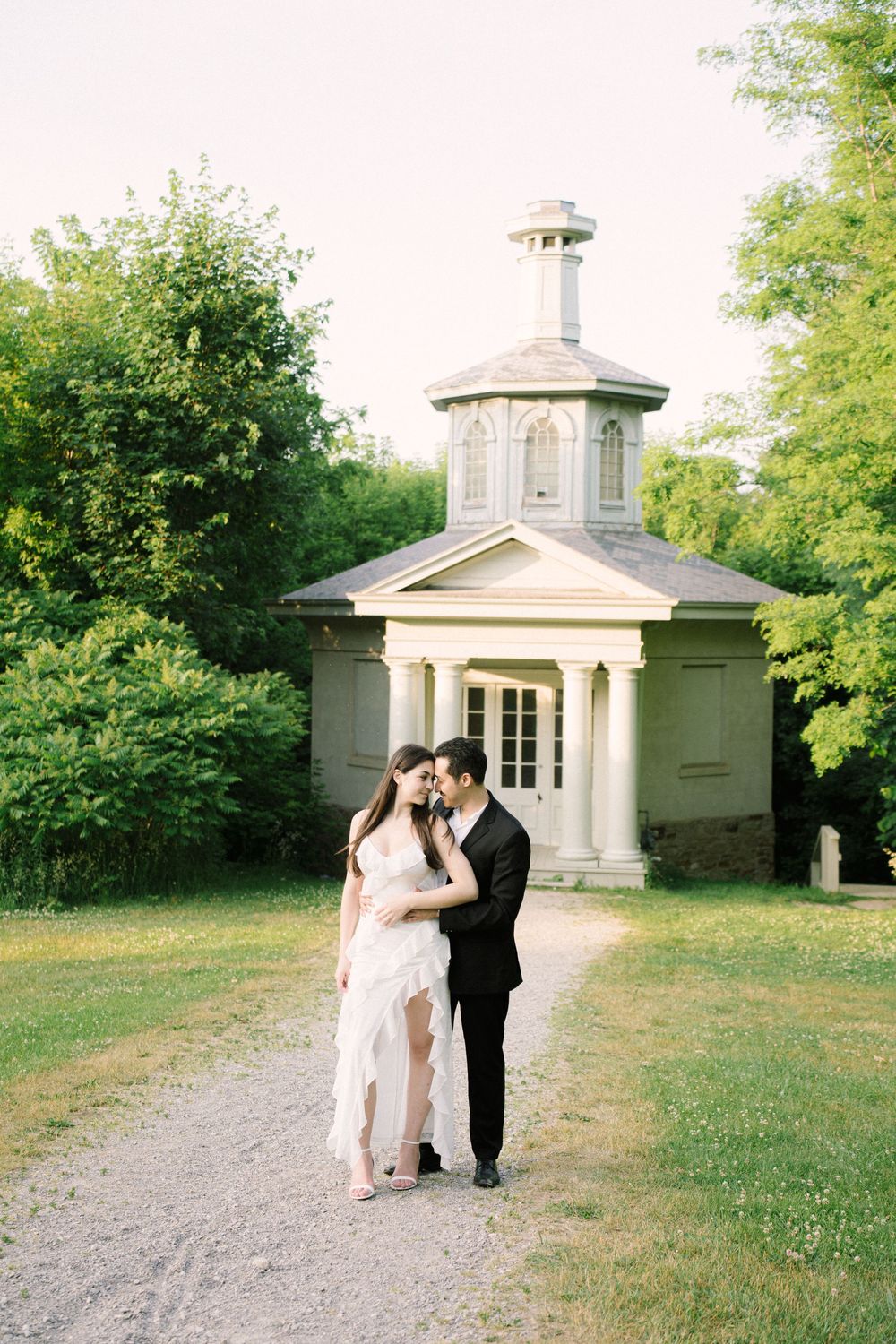 A couple shares an intimate moment in front of a small stone gazebo surrounded by lush green trees in a garden setting.