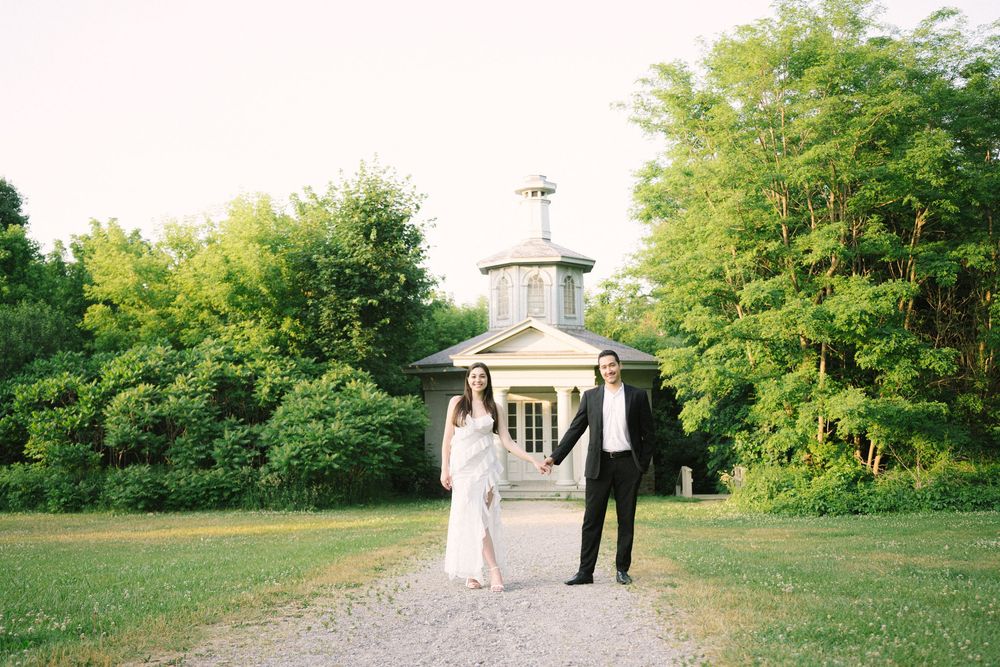 A wedding couple walks hand in hand across a scenic grassy field next to old buildings and green trees.
