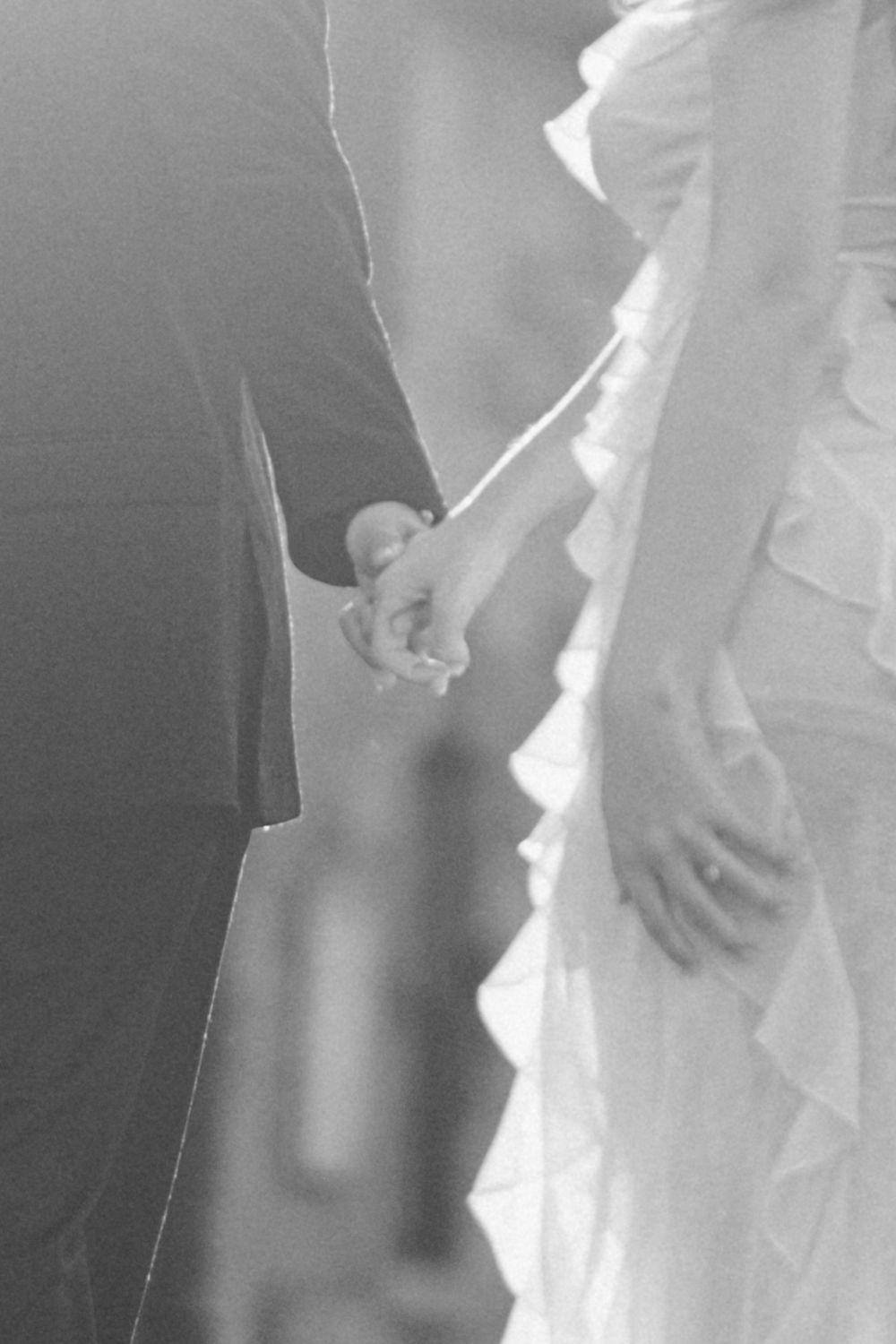 A black and white wedding photograph showing two hands clasped together, with a flowing white dress in the background.