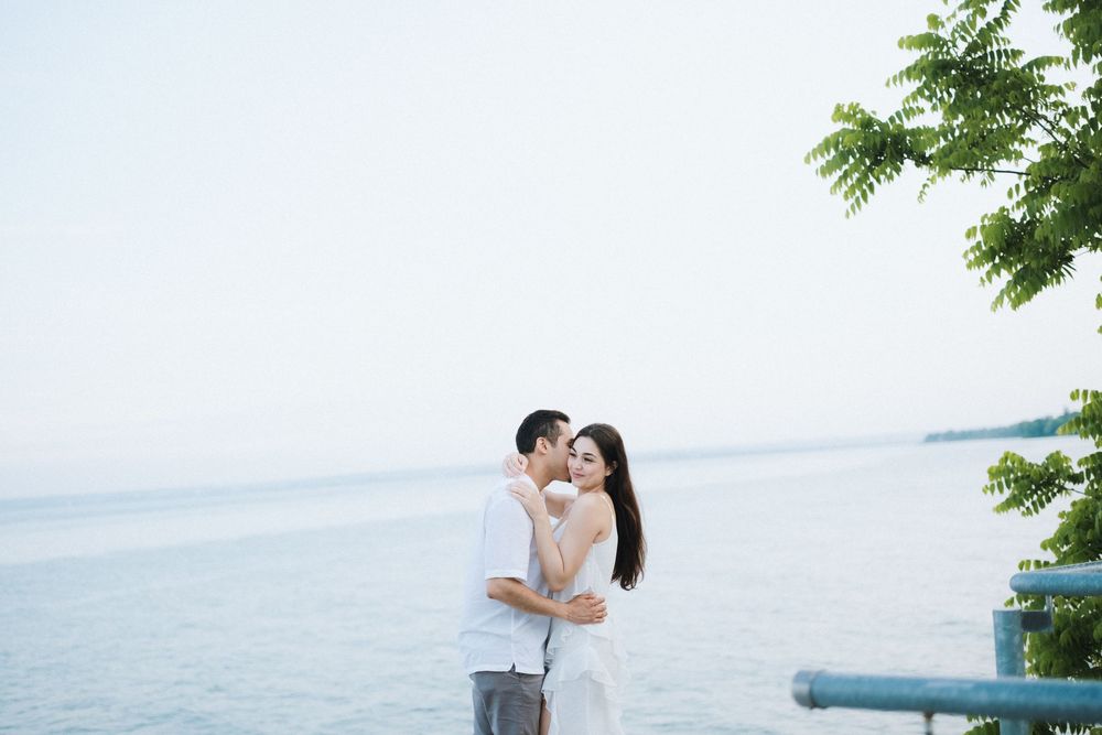 A romantic beach photoshoot showcases natural affection against a serene ocean backdrop with overhanging trees.