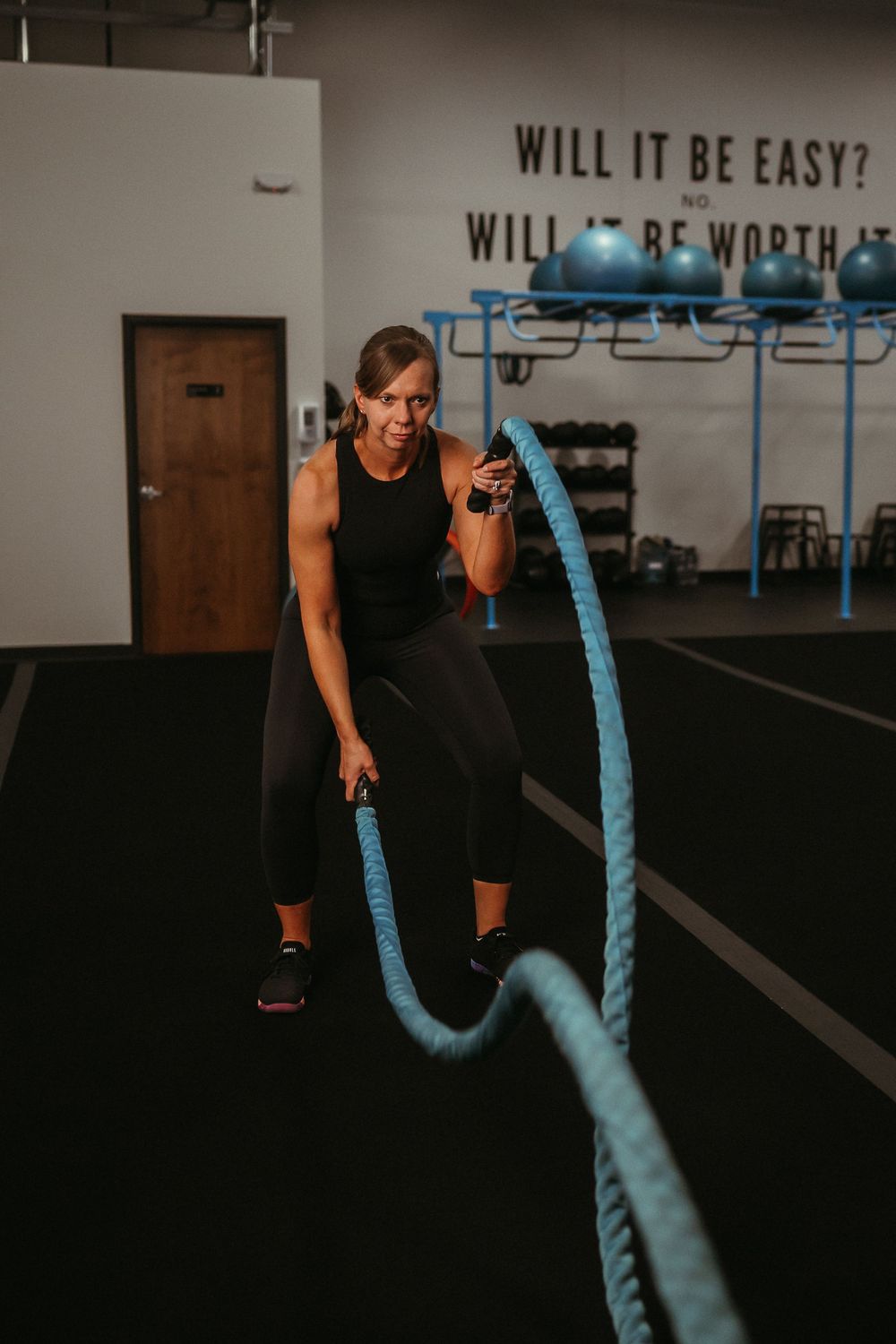 Person in black workout attire performs battle rope exercises in a dark gym setting with motivational text on walls.