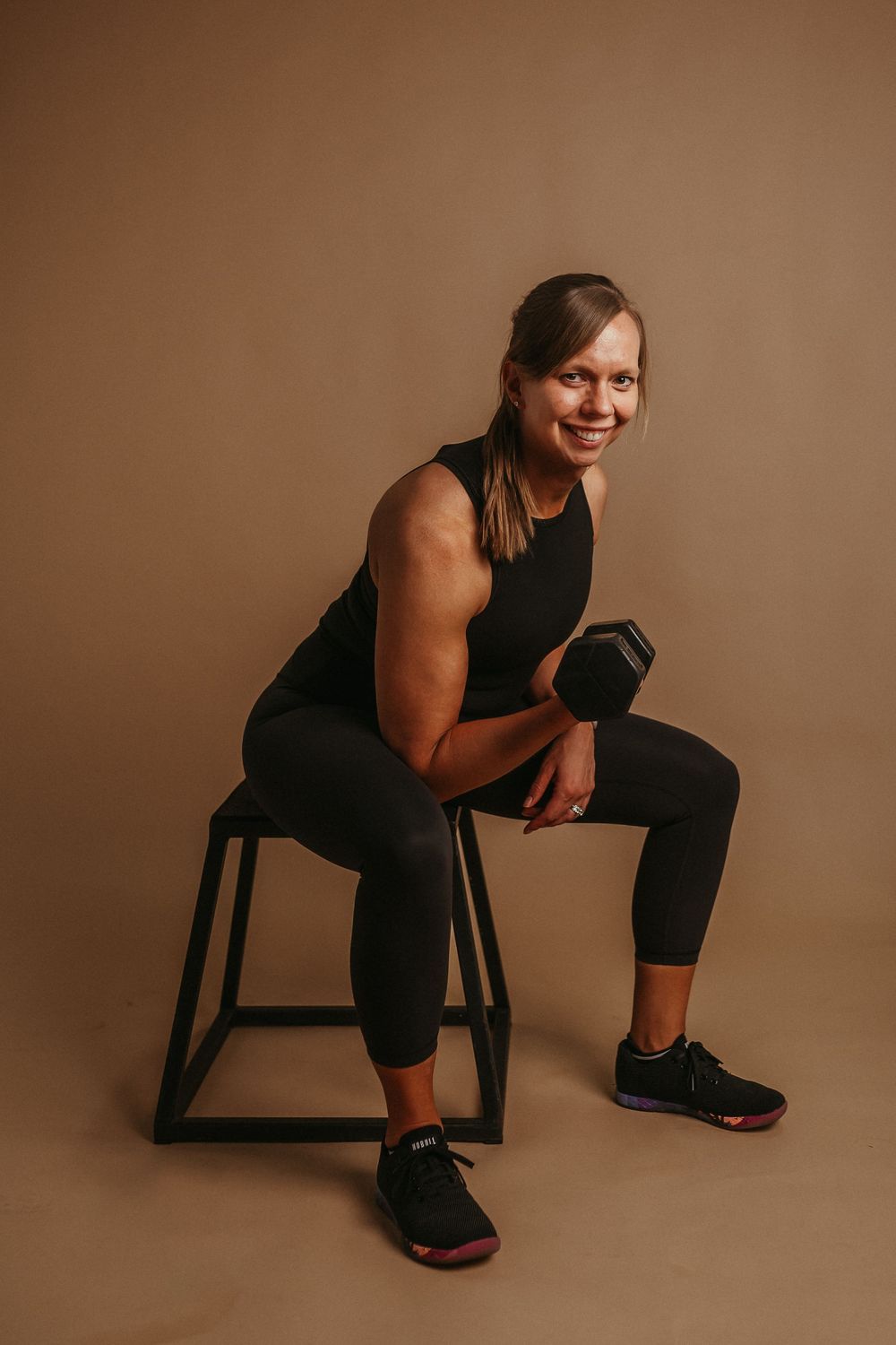 A figure in black athletic wear sits contemplatively on a wooden chair against a warm brown studio backdrop.