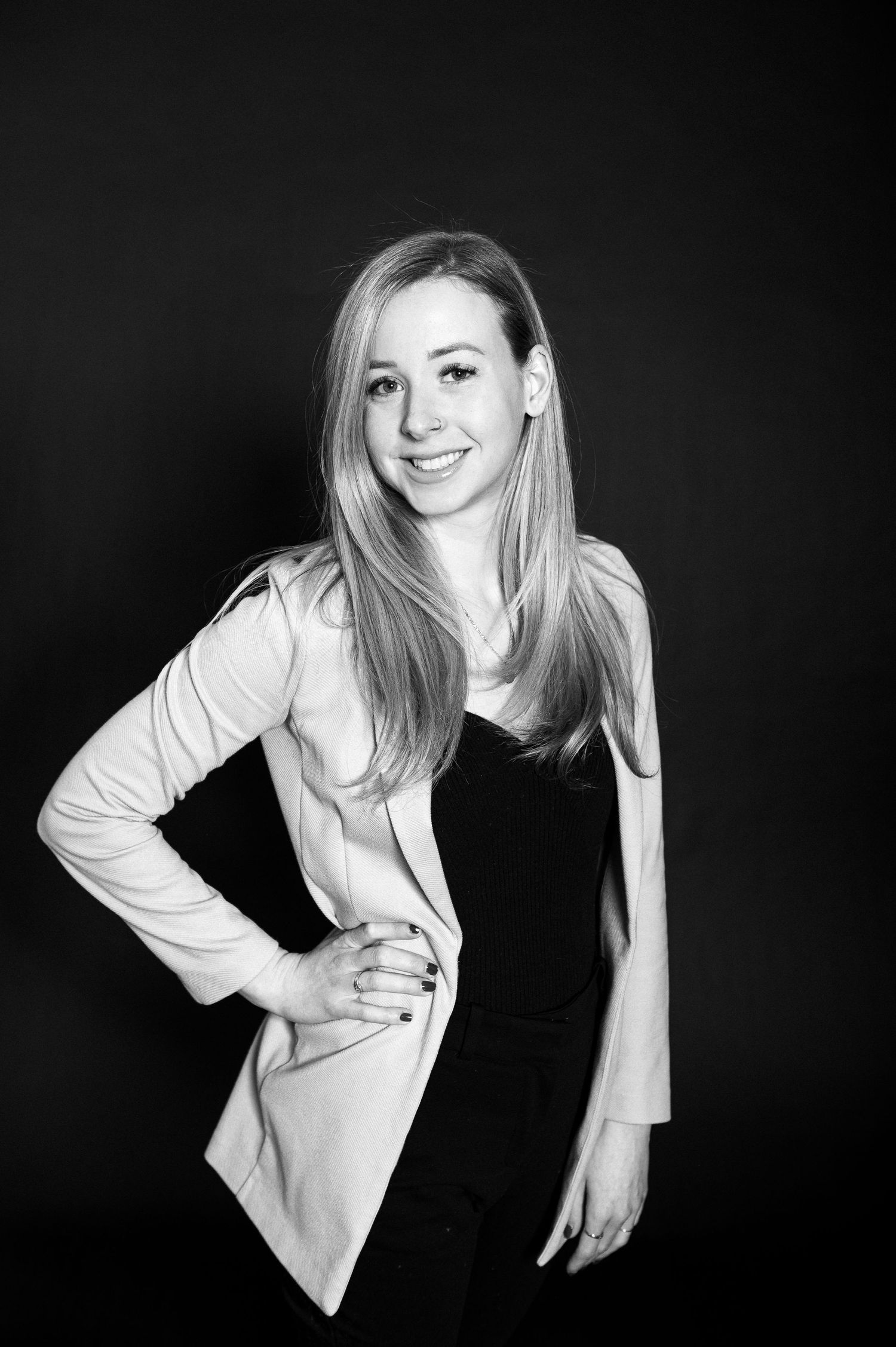 Black and white portrait of a young woman with long blonde hair wearing a light blazer over dark top against dark background.