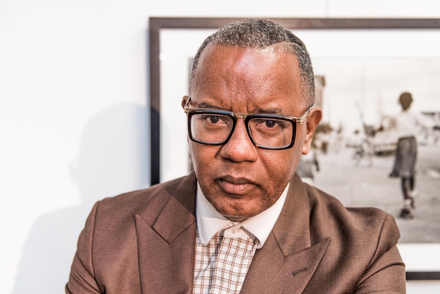 Professional in brown suit and glasses sitting in front of framed artwork.