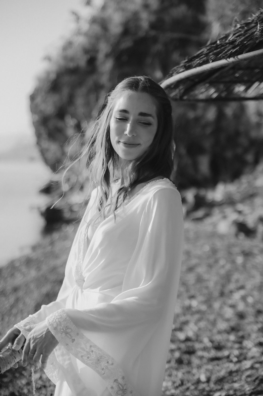 Black and white portrait of a young woman in a white flowy dress standing outdoors in natural sunlight with wind in her hair.