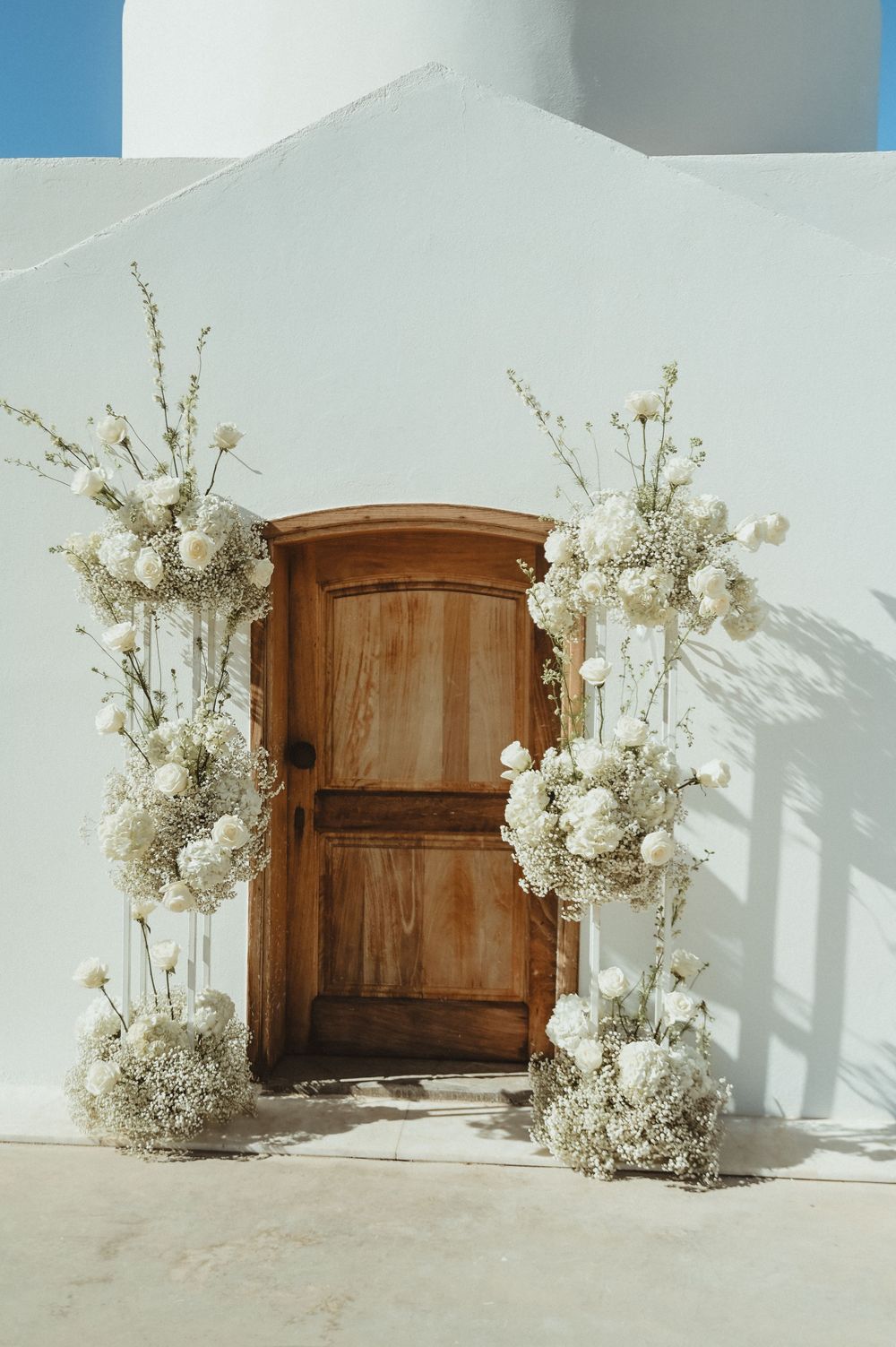 Wooden door framed by white floral arrangements against a whitewashed wall creating an elegant wedding entrance.