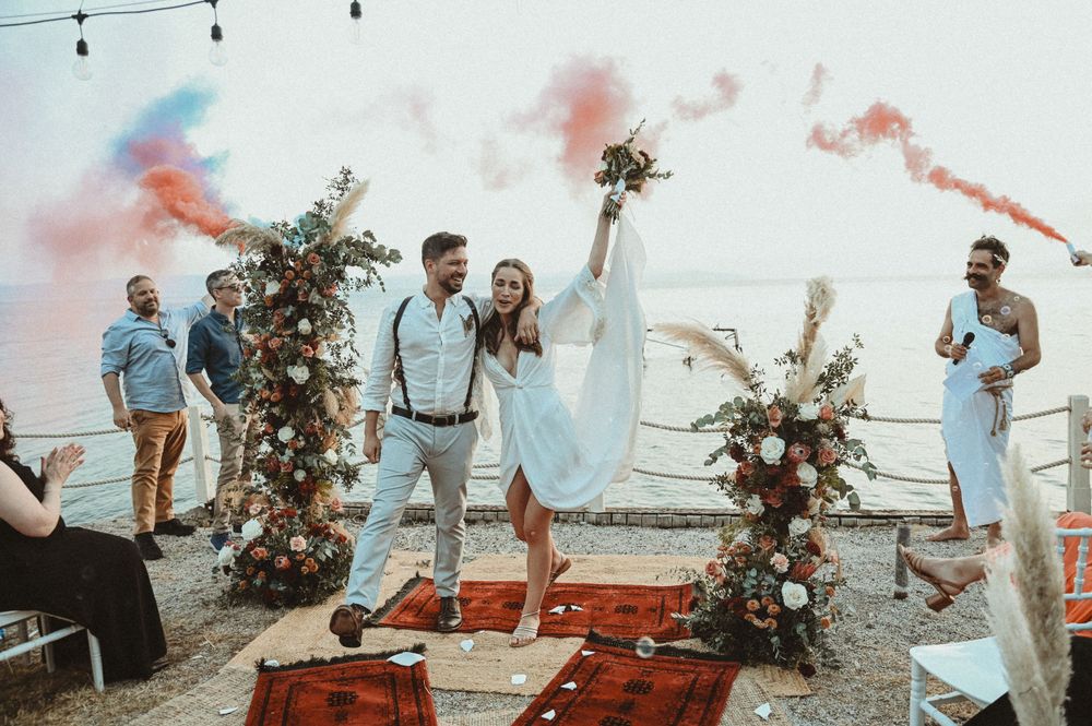 A romantic celebration at sunset with colorful smoke bombs and floral arches decorating an outdoor seaside setting.