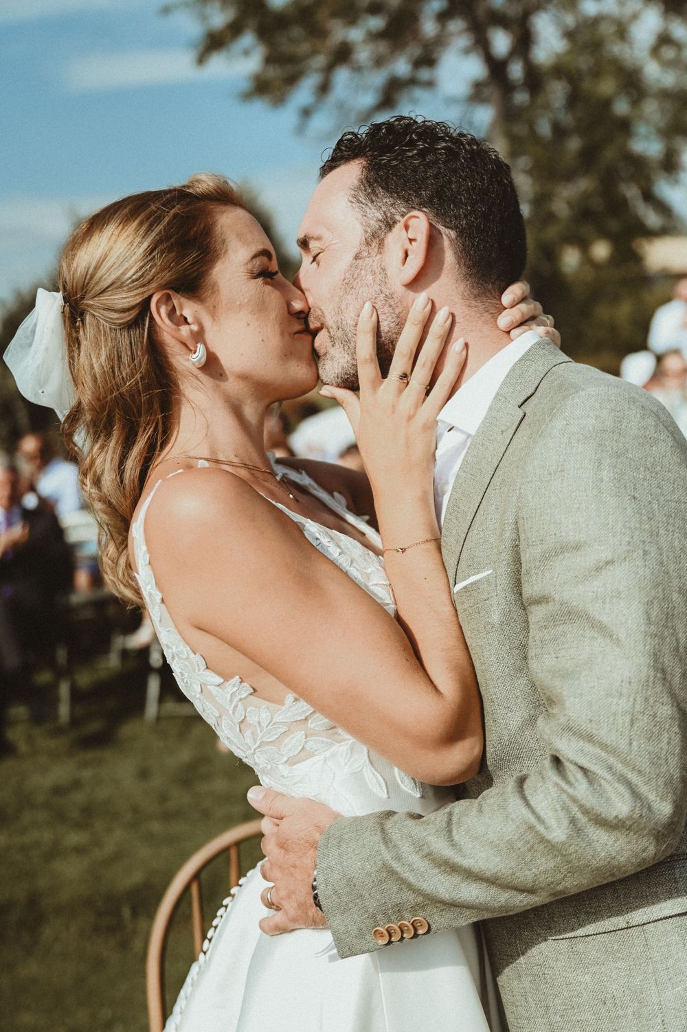 A romantic outdoor wedding kiss between newlyweds, with the bride in white and groom in light gray under blue sunny skies.