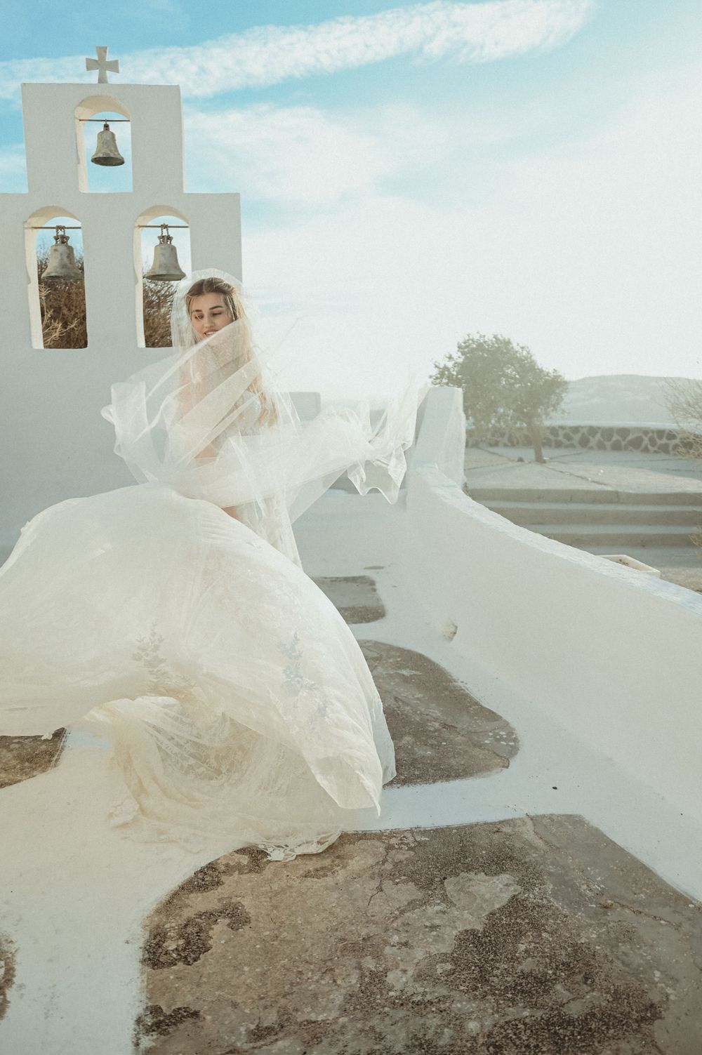 A flowing white wedding dress billows in the wind against a whitewashed Greek church with bell tower under blue skies.
