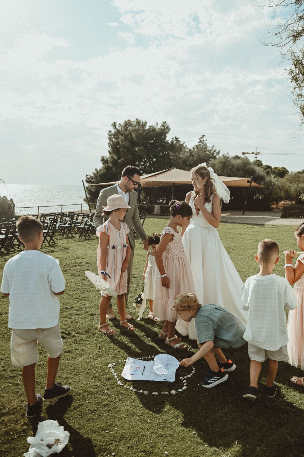A group gathers outdoors on a grassy hill during a summer wedding celebration with a scenic view in the background.